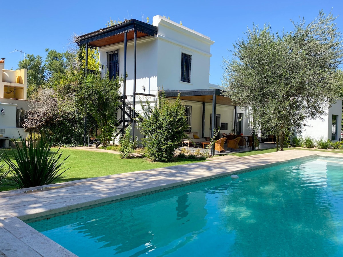 A charming property features a two-story house surrounded by a spacious garden. A shimmering pool glistens in the sunlight, while comfortable seating areas are scattered throughout the outdoor space. Lush greenery and trees enhance the tranquil setting.