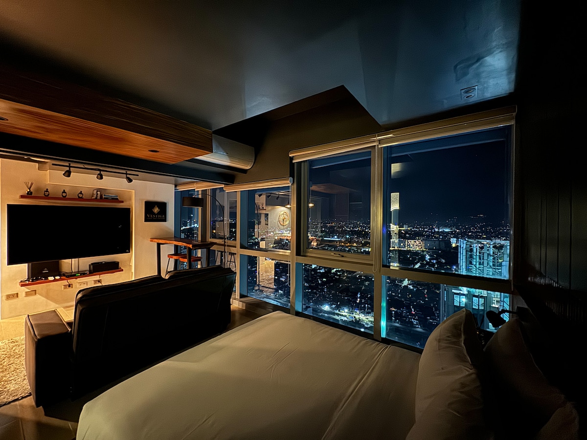 A modern industrial-themed space is showcased with panoramic floor-to-ceiling windows revealing city lights at night. A spacious bed is positioned beside a dark sofa, while a wall-mounted TV is visible in the background, contributing to the urban ambiance.