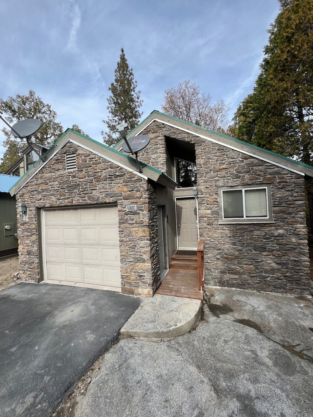 The exterior of the two-story house features a charming stone facade with a sloped roof. A small staircase leads to the front door, and a single-car garage is visible on the left. Trees surround the property, contributing to a serene forest ambiance.