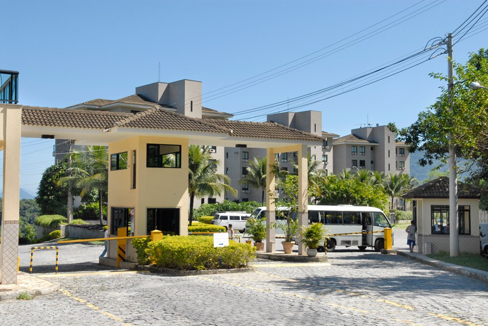 The entrance to the condominium is framed by palm trees and well-maintained gardens. A gated structure with an overhang provides access, while paved pathways extend into the community. The background features multiple buildings, showcasing the expansive area of the resort.