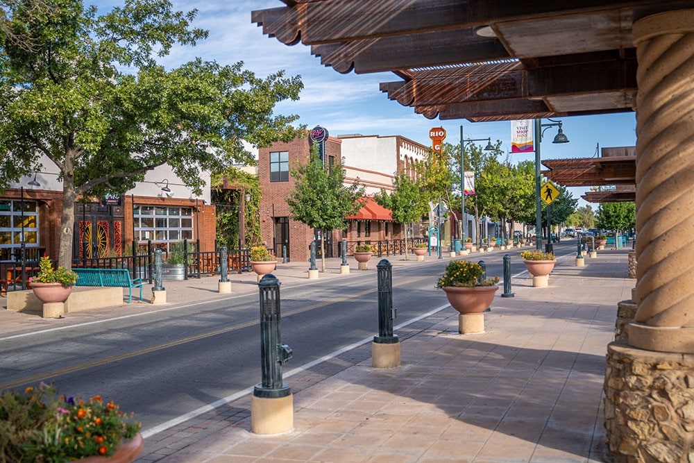 A wide street is lined with colorful buildings featuring varied architectural styles. Planters with greenery are positioned along the sidewalk, and lamps illuminate the area. Shaded seating is visible, creating a welcoming atmosphere. Clear blue skies and scattered clouds enhance the setting.