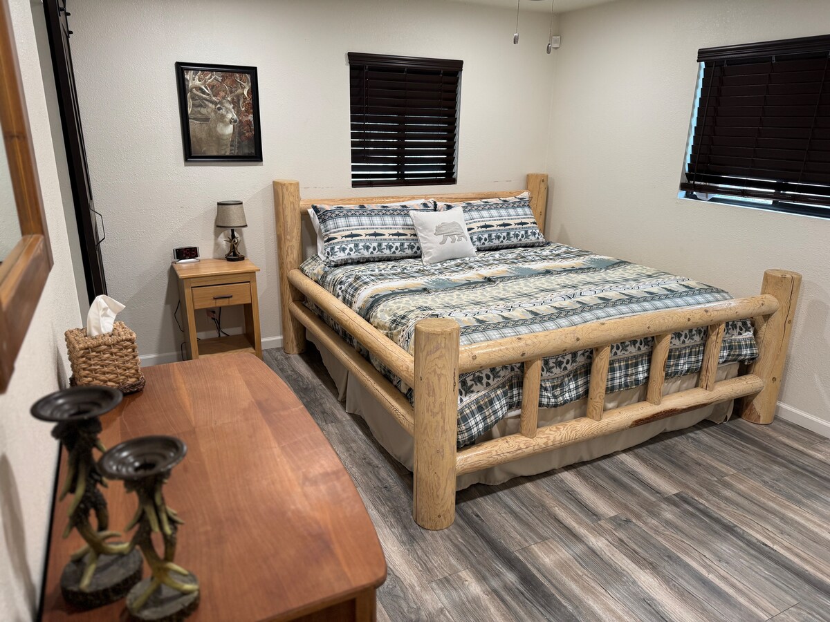A spacious sleeping area features a king-size bed with a patterned blanket draped across it. Wooden furniture, including a bedside table and a dresser, complement the log-style bed frame. Natural light enters through the window, adorned with dark blinds, creating a cozy ambiance.