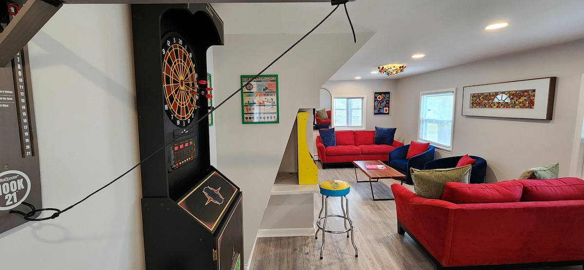 A vibrant living space features a dartboard and colorful seating, inviting relaxation and engagement. A mixture of red and yellow seating options complements the light grey walls. Natural light fills the room through multiple windows, enhancing the cheerful atmosphere.