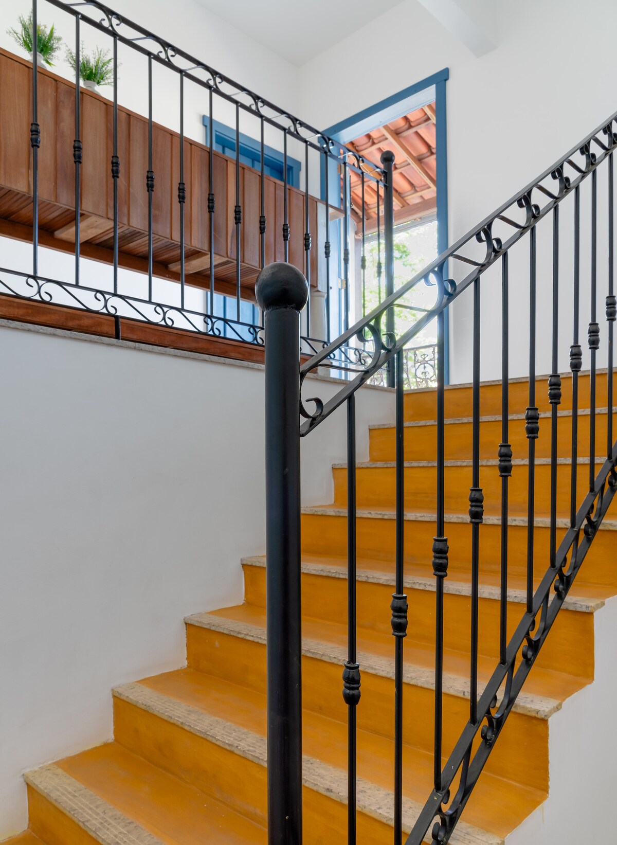 A staircase with orange painted steps leads to a light-filled upper level. The metal railing features intricate designs, providing a contrast to the bright walls. Natural light filters through a nearby window, highlighting the staircase's inviting atmosphere.