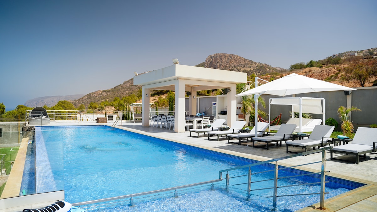 An infinity pool is visible, extending towards a stunning sea view. Lounge chairs surround the pool area, complemented by a shaded seating area under a canopy. The backdrop features rolling hills and clear blue skies, creating a serene outdoor environment.