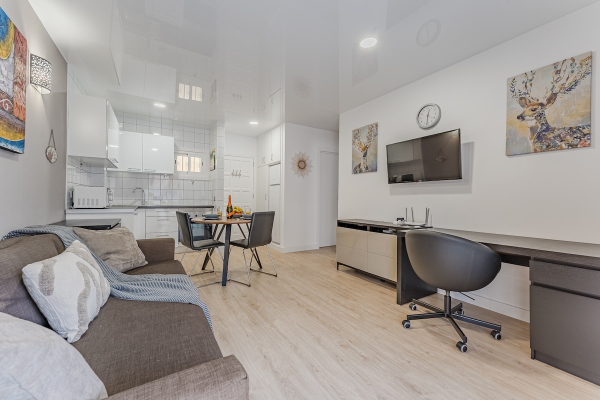 A modern living space features a cozy sofa with decorative pillows, a round dining table with seating for four, and a workspace with a black office chair. The kitchen area includes sleek cabinetry and appliances, while wall art and a clock add visual interest.