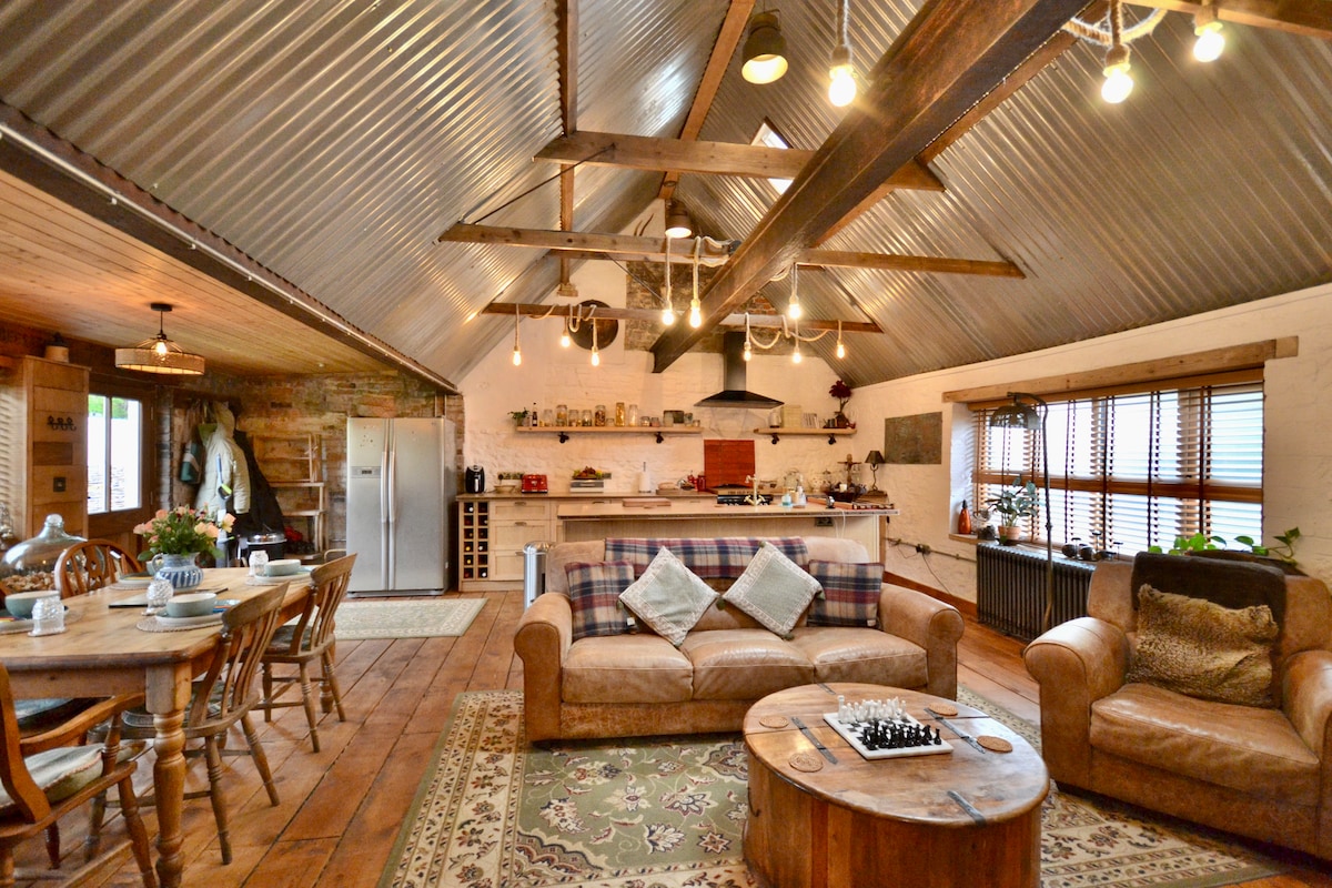 An open-plan living area features a rustic design with a high, corrugated metal ceiling and wooden beams. A large, inviting sofa is centered around a round coffee table with a chess set. A dining table is set with dinnerware, and a well-equipped kitchen is visible in the background.