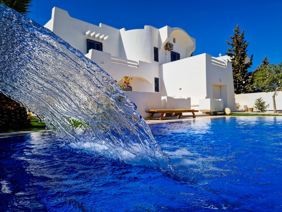 The image features a sparkling blue swimming pool with water cascading from a fountain, creating a serene effect. In the background, a white villa is visible, surrounded by lush greenery and clear skies, enhancing the tranquil outdoor space.