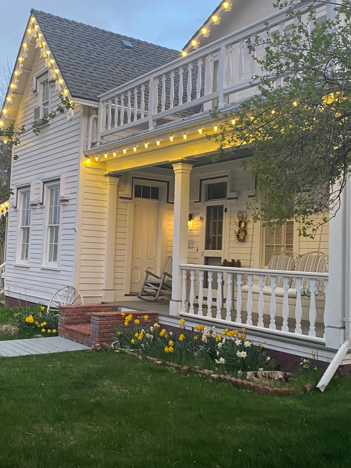 A charming exterior view of a two-story house is presented, featuring a wraparound porch adorned with white rocking chairs. Soft yellow string lights illuminate the porch and garden area, where vibrant yellow tulips bloom against a backdrop of lush green grass.