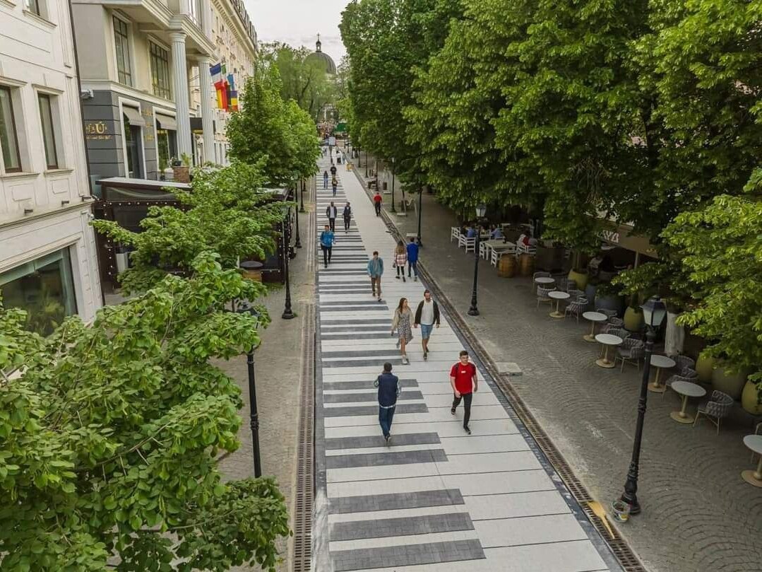 A vibrant pedestrian street is lined with lush trees, providing shade to visitors strolling along. A large, artistic piano design spans the pavement, while cafes and restaurants are visible on either side, enhancing the lively atmosphere of the area.
