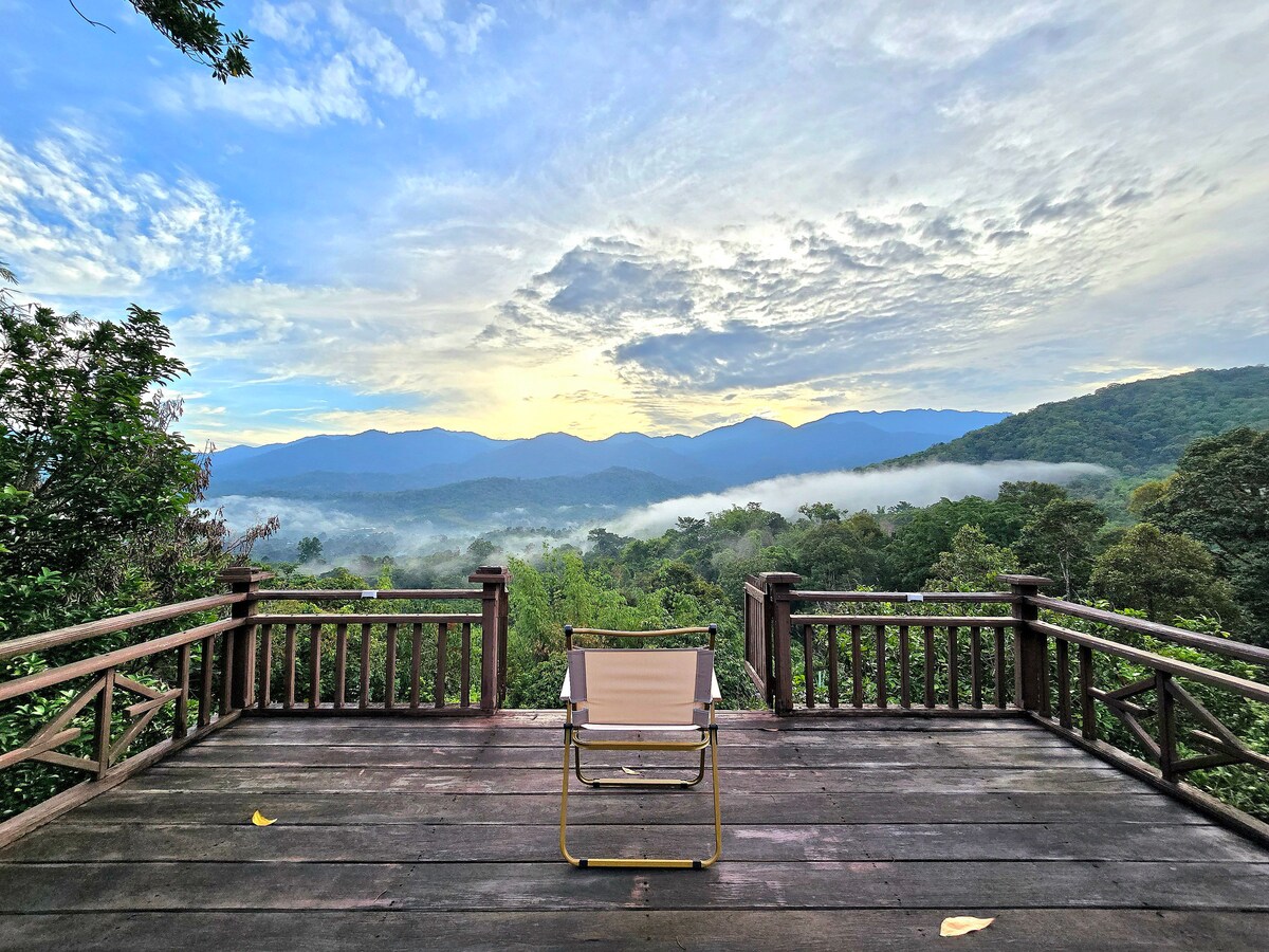 A spacious viewing deck is shown, featuring a single chair facing a stunning panoramic vista of mountains and lush greenery. The sky displays a range of colors as clouds drift, creating a serene backdrop for a tranquil outdoor experience.