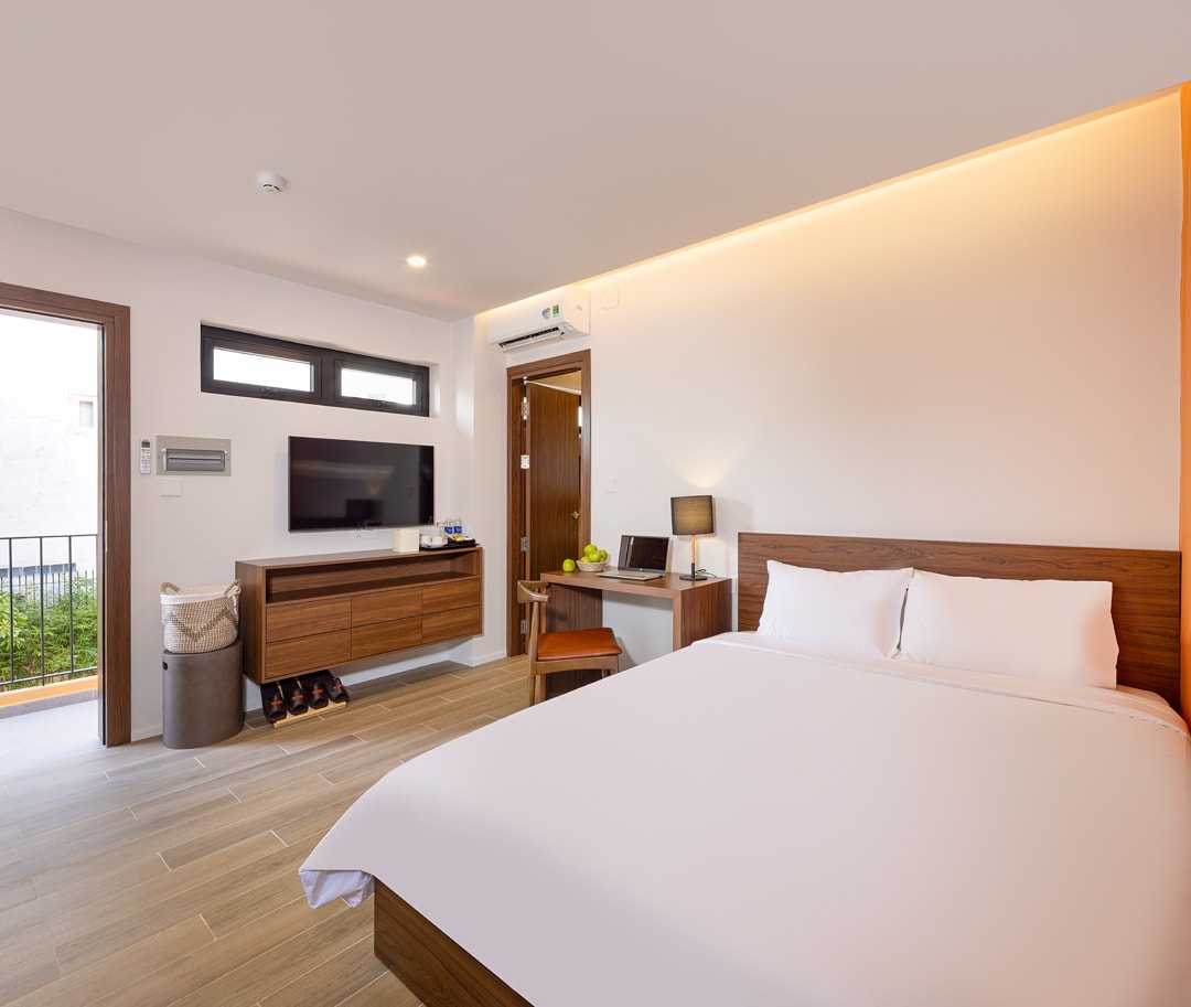 A room features a spacious bed with crisp white linens, a wooden dresser beneath a flat-screen TV, and a small desk with a chair. Natural light enters through large windows, while a door leads to a private outdoor area with greenery.