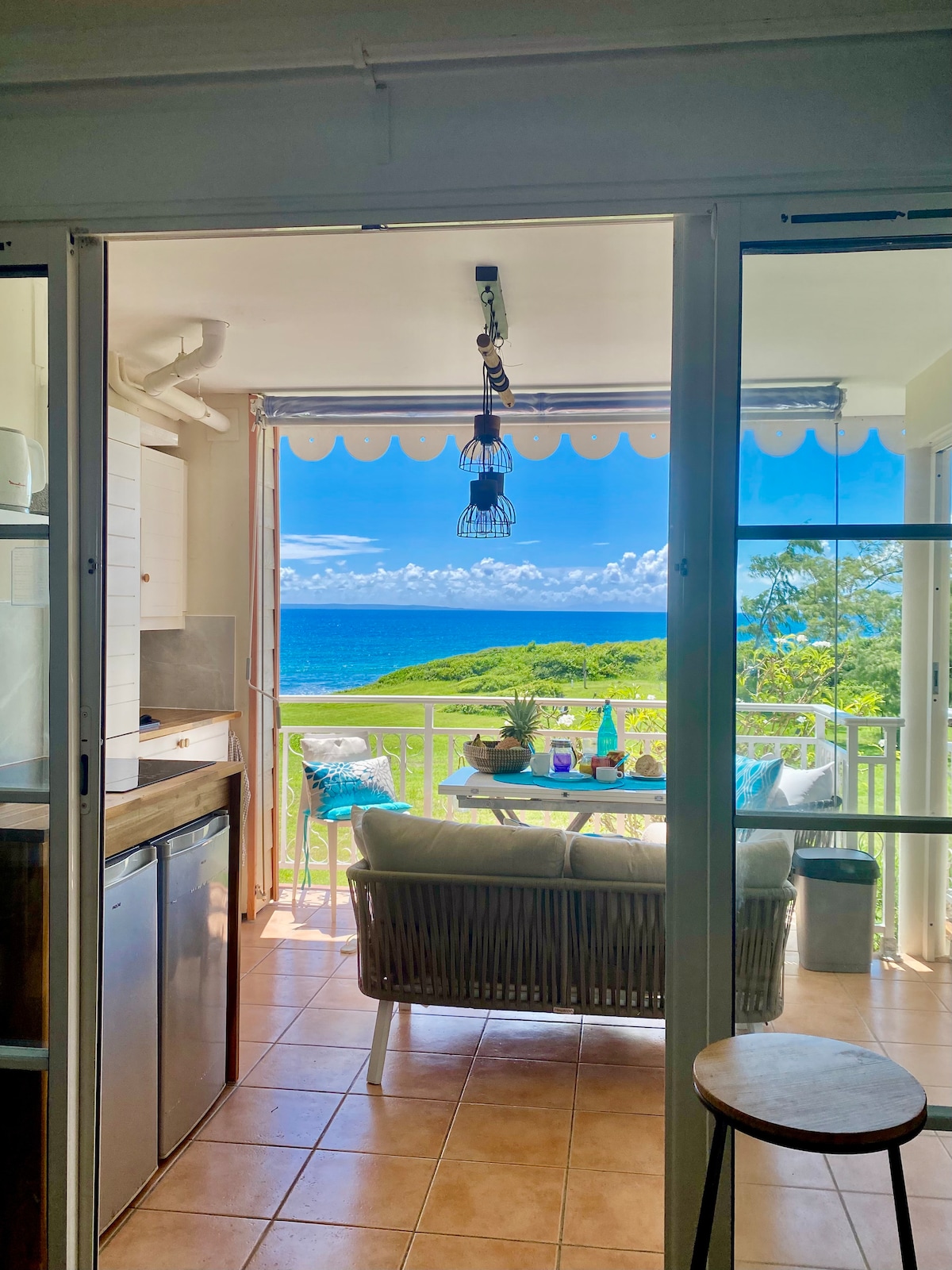 A bright and airy indoor space features a glimpse of the terrace, with ocean views visible through glass doors. A comfortable outdoor seating area is set up with a small table, inviting relaxation while enjoying the surrounding landscape. Natural light enhances the tranquil atmosphere.