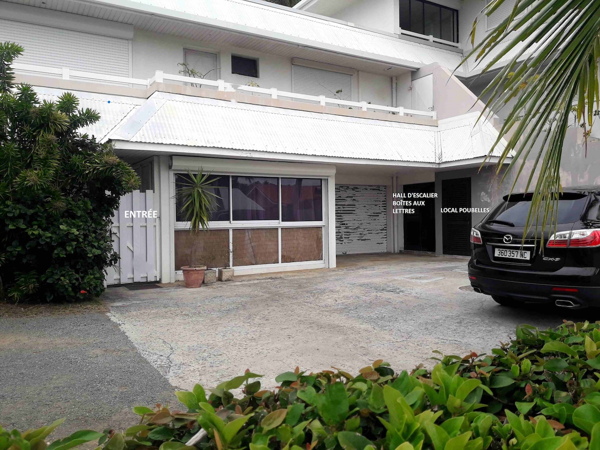 A driveway leads to the front of the property, featuring an entrance gate and a spacious carport. The façade includes large windows that allow for natural light, while greenery borders the area, providing a sense of privacy. Outdoor space is paved for easy access.