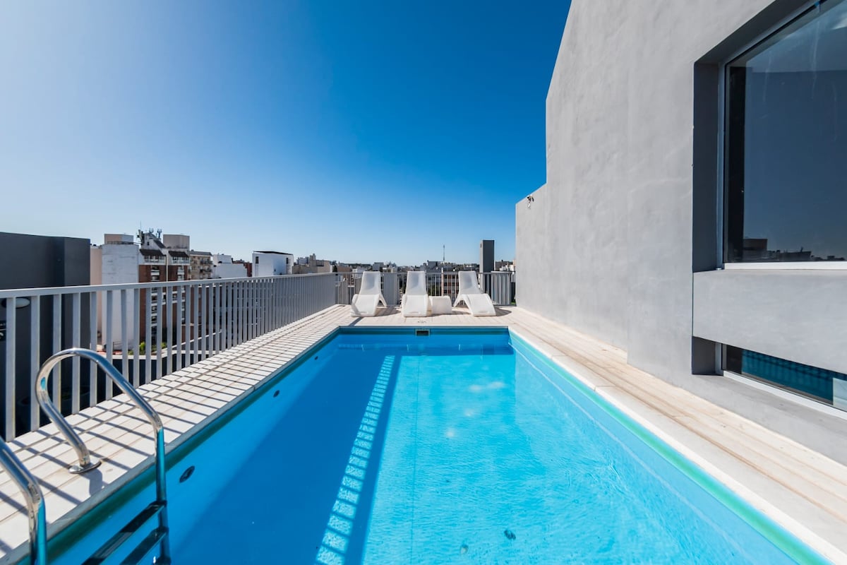 A rooftop swimming pool is visible under a clear blue sky, with a sun deck featuring several lounge chairs nearby. The pool's shimmering water reflects the sunlight, creating a serene atmosphere. The surrounding skyline is partially shown, hinting at urban views.