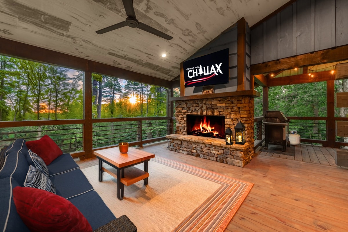 A spacious outdoor living area is highlighted by a stone fireplace, complemented by a plush sectional and decorative pillows. A wooden coffee table rests on a large area rug. A grill is positioned nearby, and large windows showcase views of the surrounding trees.