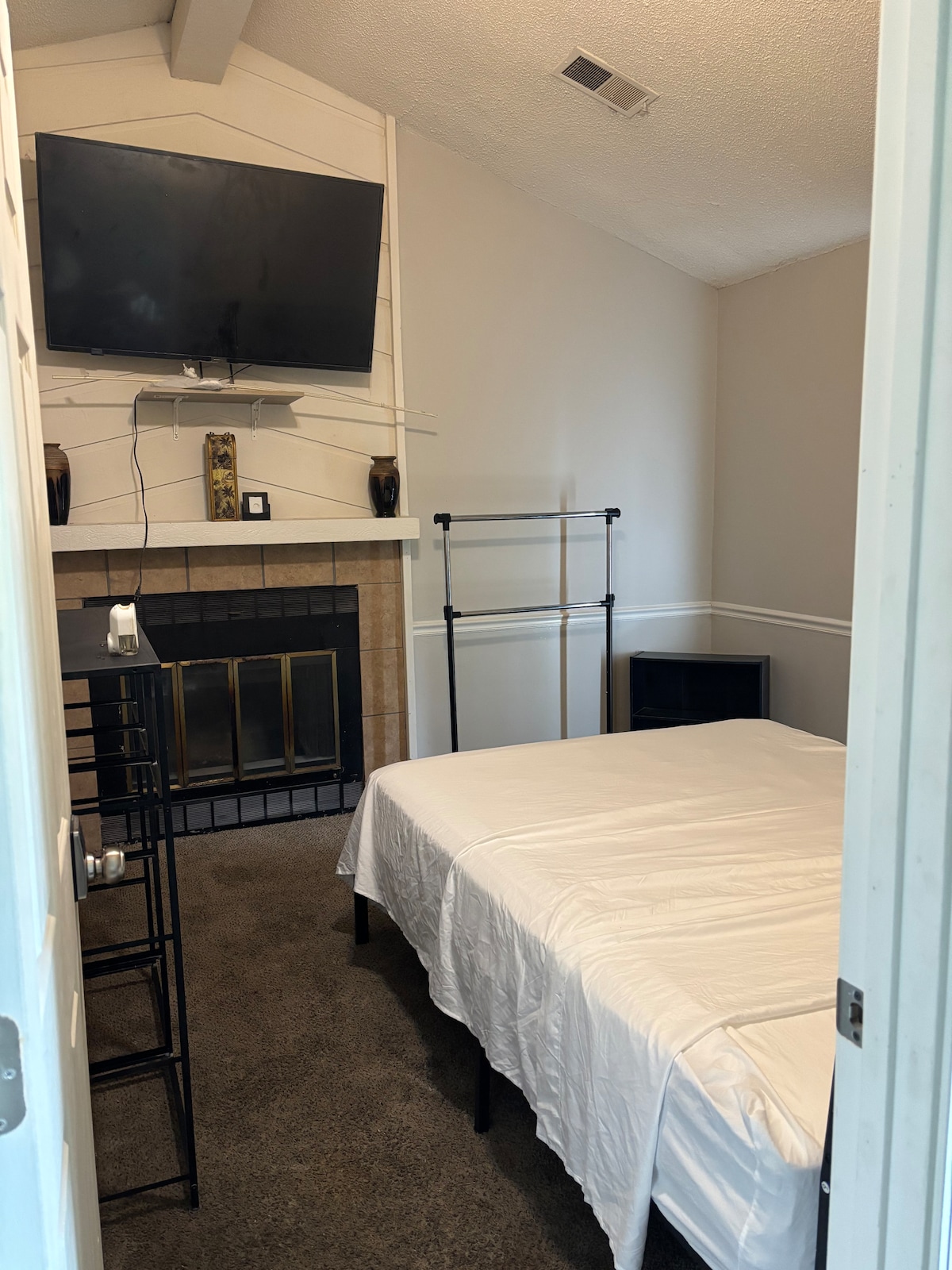 A well-lit room features a twin size bed with a white bedspread, positioned next to a black nightstand. A television is mounted above a fireplace against a shiplap wall. An adjustable clothing rack is visible, adding functionality to the space.