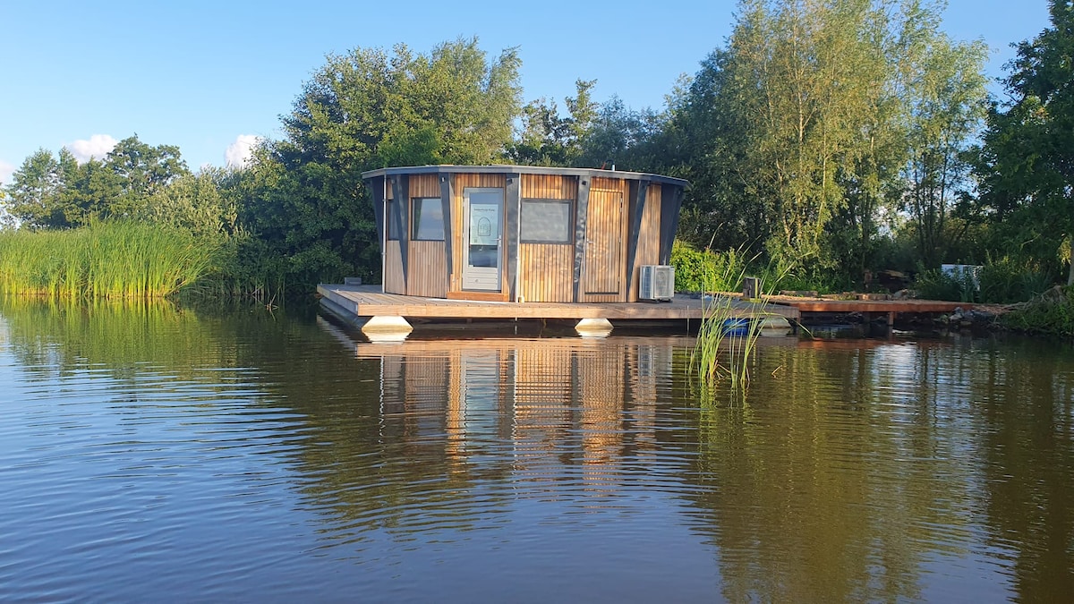 A compact house is situated on stilts over calm water, featuring a circular design with wooden paneling. Surrounding vegetation includes lush reeds and trees, contributing to a serene outdoor environment. A wooden deck extends from the house, offering access to the water.