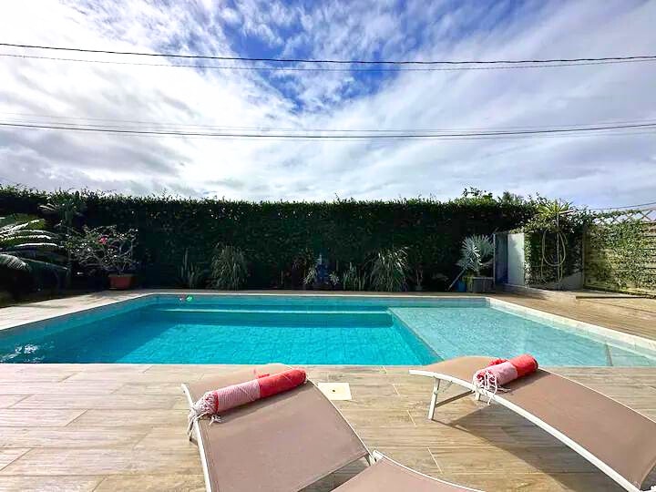 A clear swimming pool is surrounded by a wooden deck, with two lounge chairs positioned nearby. Lush greenery and flowering plants are visible in the background, complemented by a partly cloudy sky overhead.