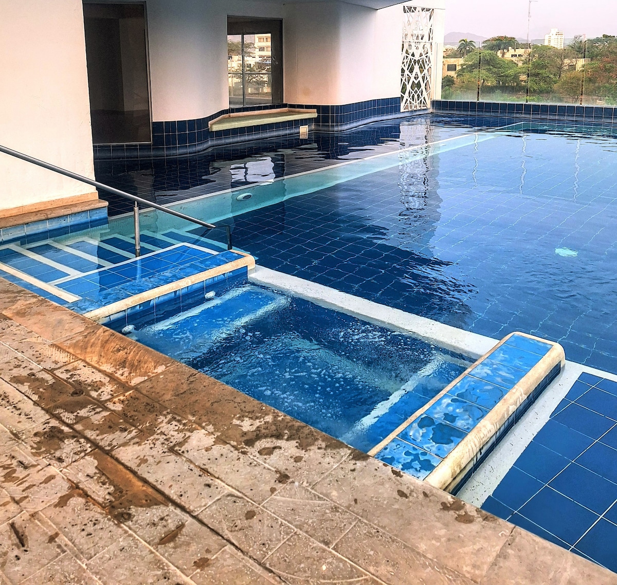 A swimming pool area features clear water with a modern design, including a shallow section and contrasting blue tiles. Sunlight reflects off the surface, while steps lead from the pool to a nearby seating area. Surrounding greenery adds to the serene atmosphere.