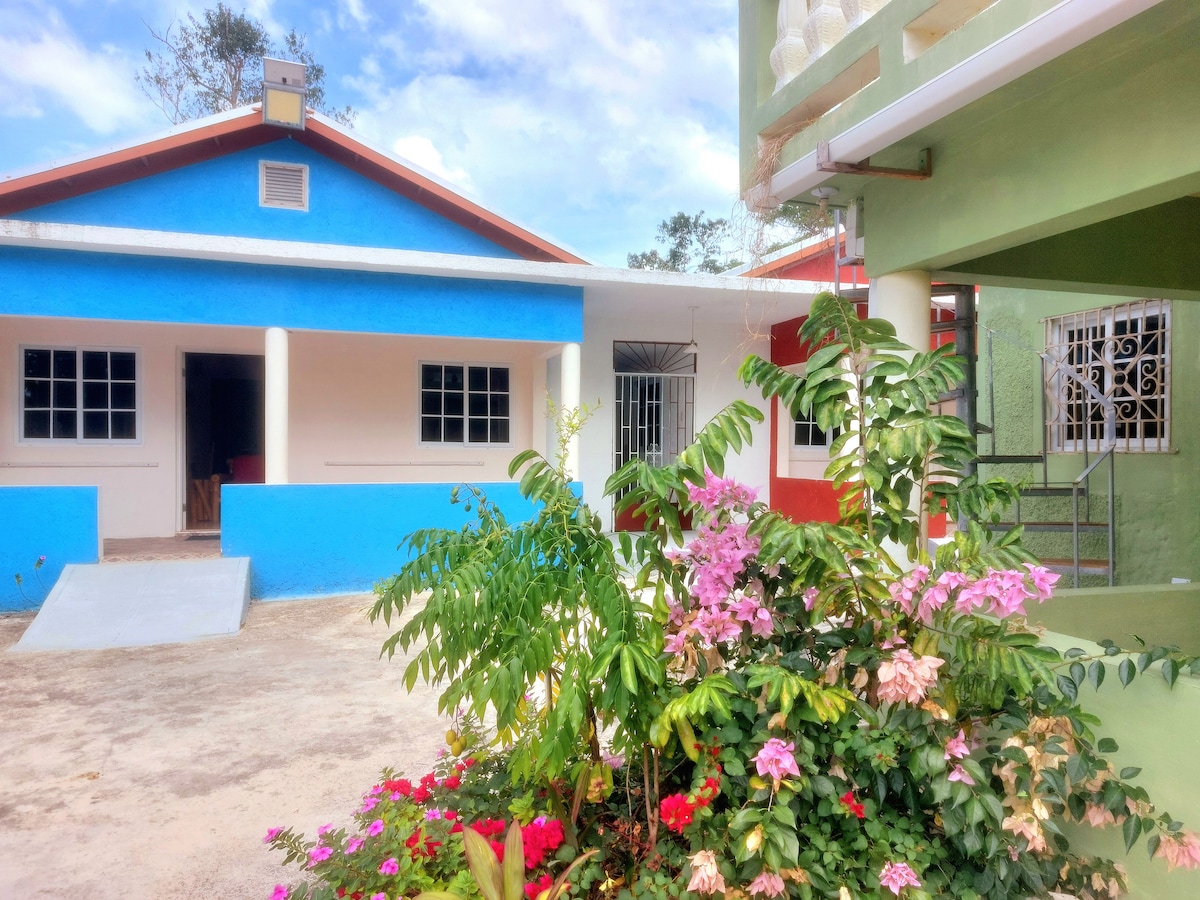 An exterior view of Stoney Gate Cottages features bright building facades in blue, red, and cream. Lush greenery and flowering plants add vibrant color to the courtyard, while a ramp provides wheelchair access. The scene captures a welcoming atmosphere, ideal for relaxation and exploration.
