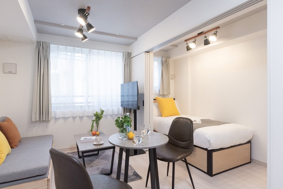 A modern living space is visible, featuring a cozy sleeping area with a spacious bed draped in white bedding. Natural light floods through large windows framed by soft curtains. A small dining table sits in the foreground, accompanied by two chairs and a decorative centerpiece of fresh fruits.