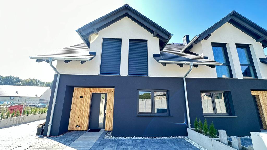 A modern two-story building features a striking combination of dark and light exterior colors. Large windows allow natural light to fill the space, while the entrance is framed by a sleek door and surrounded by neatly landscaped paths and greenery.