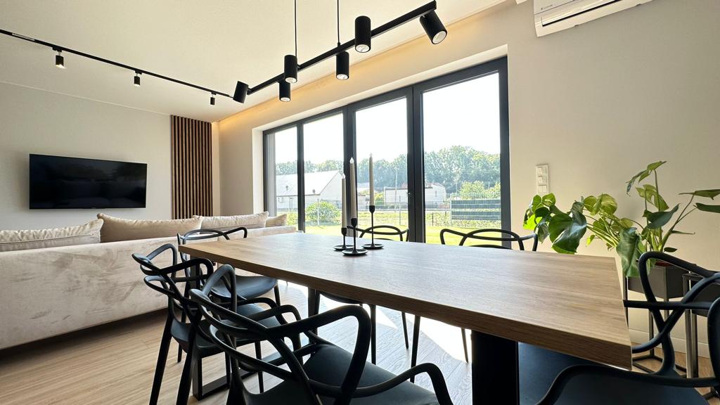 A modern dining area showcases a long wooden table surrounded by black chairs. Large windows provide natural light and views of greenery outside, while a sleek television is mounted on the wall. Subtle lighting fixtures enhance the space's contemporary feel.