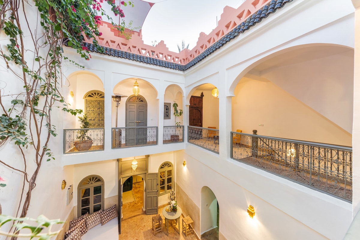 An open courtyard within the Riad is surrounded by elegantly arched doorways, showcasing intricate wrought iron railings. Natural light fills the space, illuminating the decorative features and potted greenery, contributing to a serene ambiance. Tables and chairs are arranged for relaxation.
