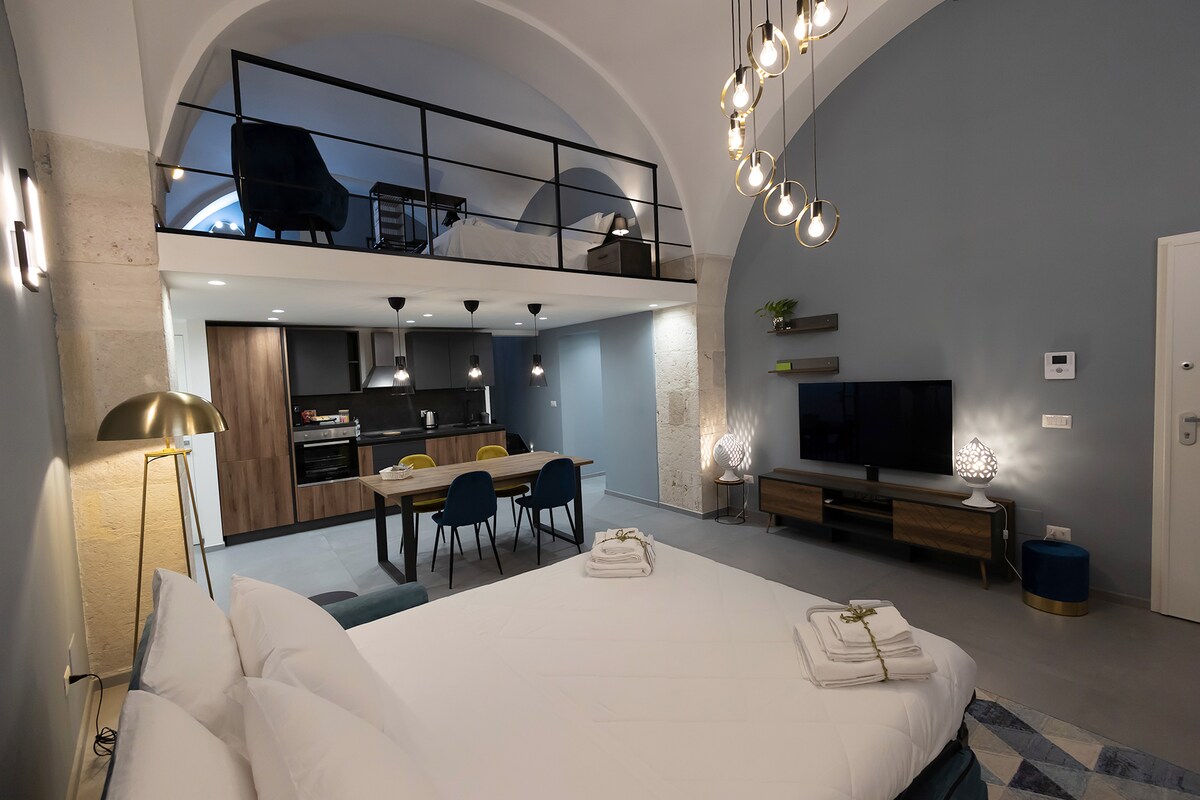 An open layout apartment features a spacious sleeping area with a large bed, modern light fixtures, and high ceilings. A dining table with chairs is positioned near the kitchen, which is equipped with contemporary appliances. A TV unit sits opposite the seating area, creating a cozy living space.