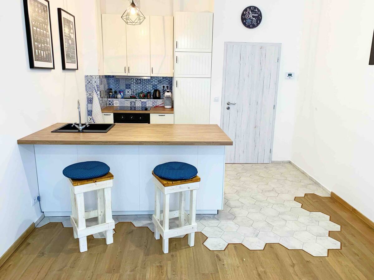 A modern kitchen area is highlighted by a wood-topped island with two wooden stools. The cabinets are white, complemented by blue tile backsplash behind the sink and stove. An inviting hexagonal floor pattern adds visual interest, with a simple door visible in the background.