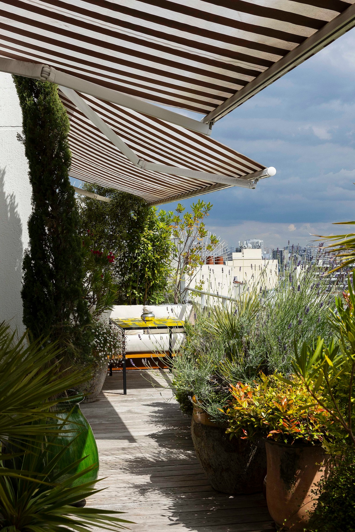 A terrace features striped awnings providing shade over a seating area. Potted plants, including small shrubs and flowering species, add greenery, while wooden decking leads to expansive views of the surrounding cityscape.