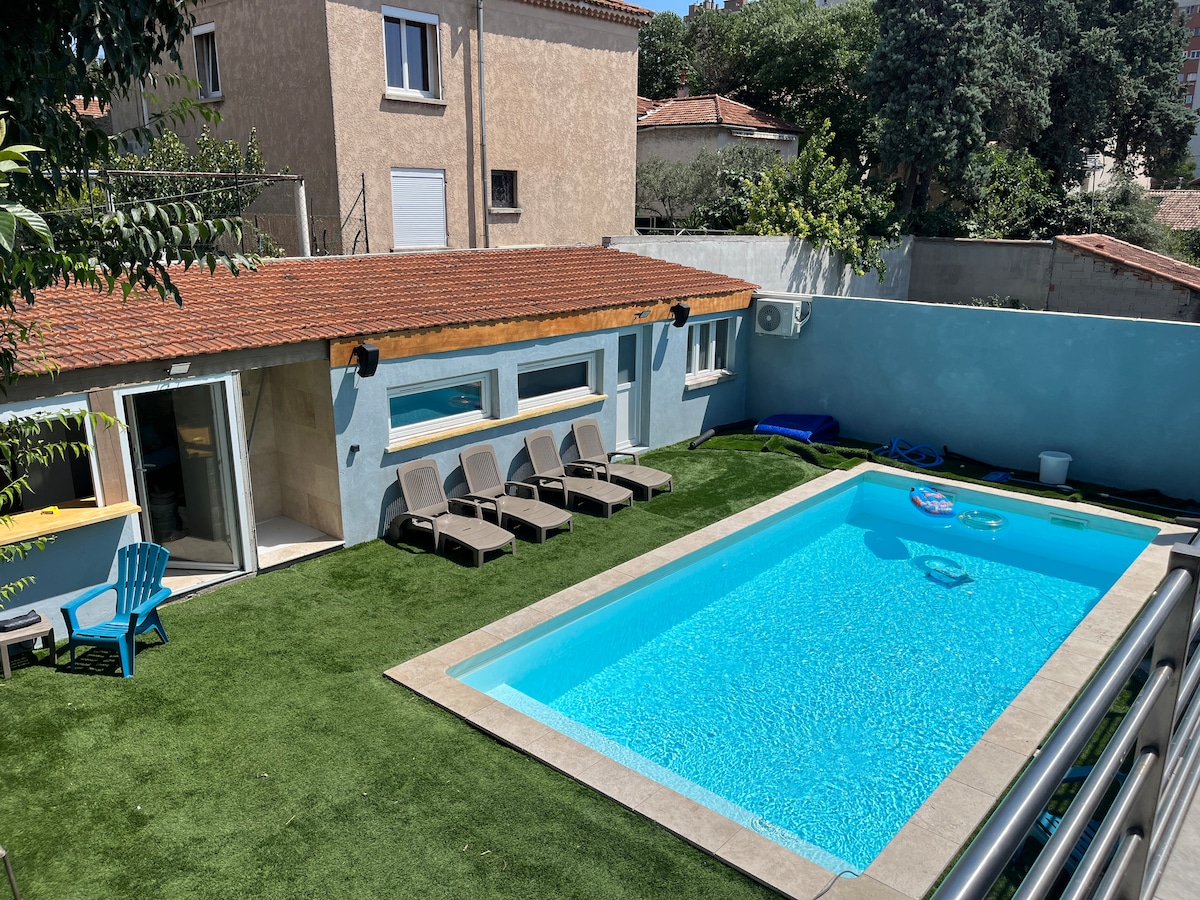 A refreshing pool is surrounded by a neatly manicured lawn, featuring several lounge chairs positioned nearby. The outdoor area includes a patio with sliding glass doors leading into a light-filled space. The backdrop includes a building with muted tones and greenery adding to the serene setting.