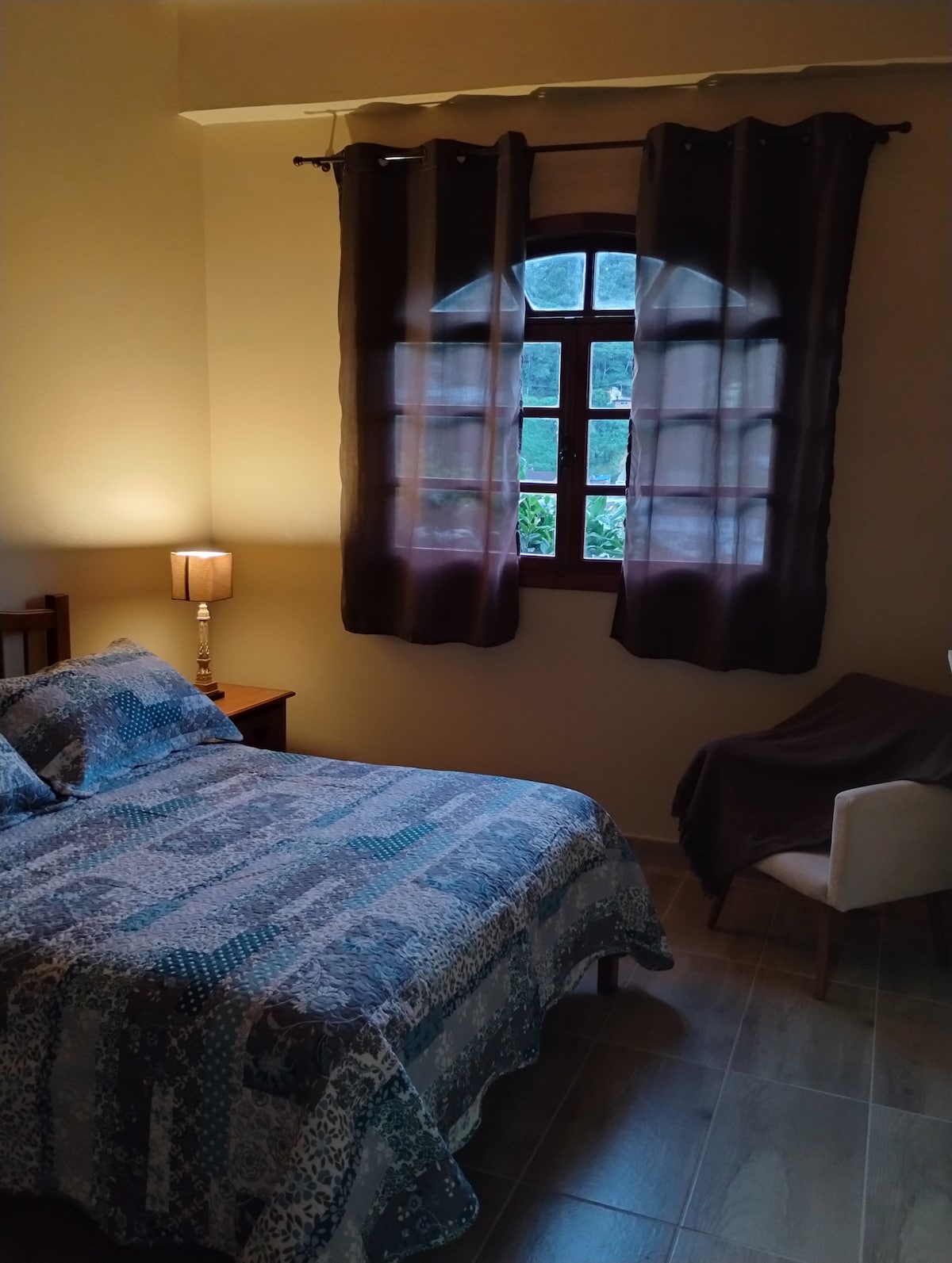 A cozy sleeping area features a patterned bedspread over a spacious bed. Light from a lamp softly illuminates the room, complemented by a window adorned with dark curtains. A small chair is positioned nearby, creating a comfortable nook.