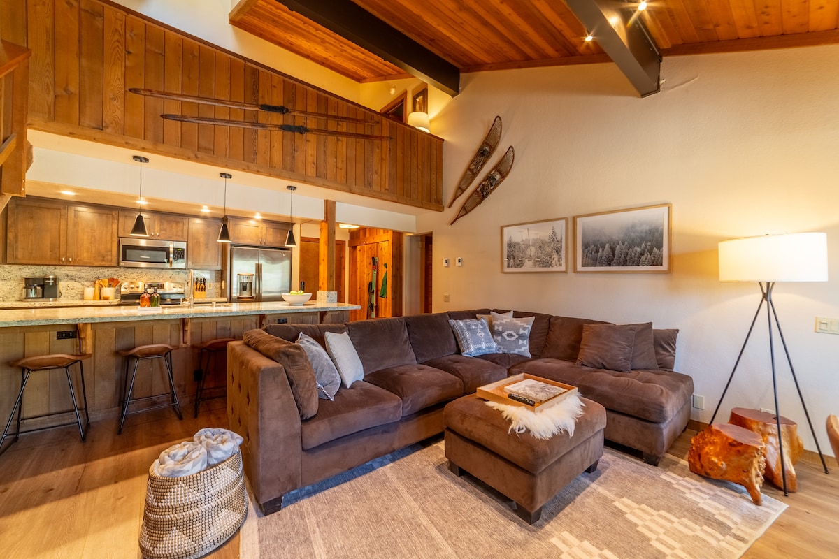 A spacious living area features a large sectional sofa inviting relaxation. Warm wood accents complement the neutral tones, while modern kitchen appliances are visible in the background. Ski decor hangs on the wall, and a cozy light illuminates the area, creating an inviting space for gatherings.