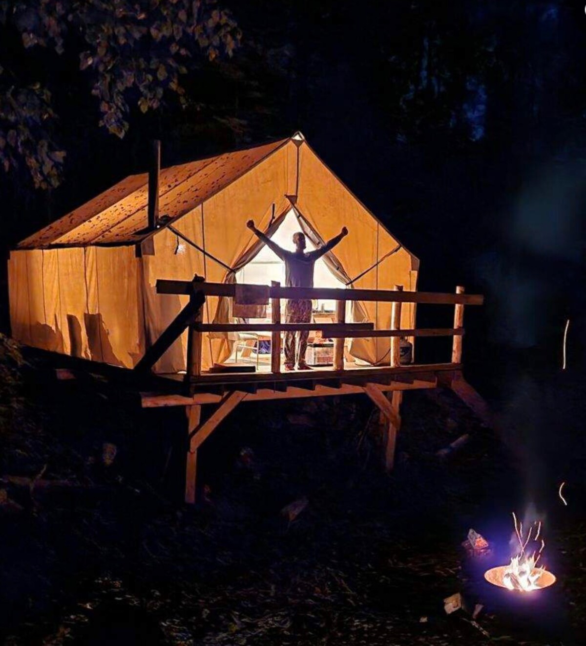 A well-lit canvas tent is positioned on a wooden deck, surrounded by the dark wilderness. The warm glow from inside the tent highlights its entrance, while a campfire burns nearby, creating a cozy atmosphere. A silhouetted figure stands with arms raised, embracing the experience.
