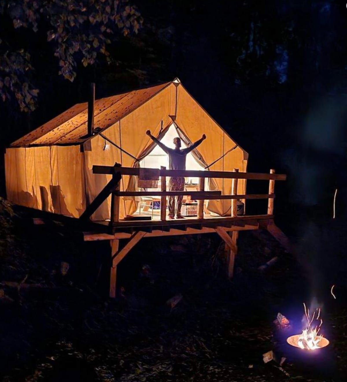 A canvas cabin tent stands on a raised deck, illuminated from within as night falls. A campfire crackles nearby, casting a warm glow. The silhouette of a person is seen with arms raised in celebration, embracing the surrounding wilderness.