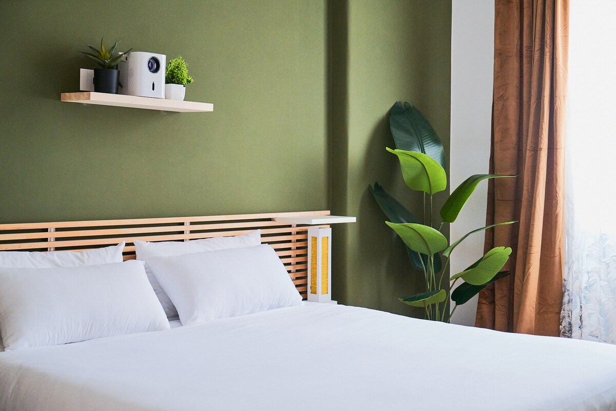 A spacious bedroom features a simple wooden headboard against a green wall. Two pillows are placed on a large white bed. A shelf above holds decorative plants, while a potted leaf plant stands beside the bed. Natural light filters in through the window, adorned with soft curtains.