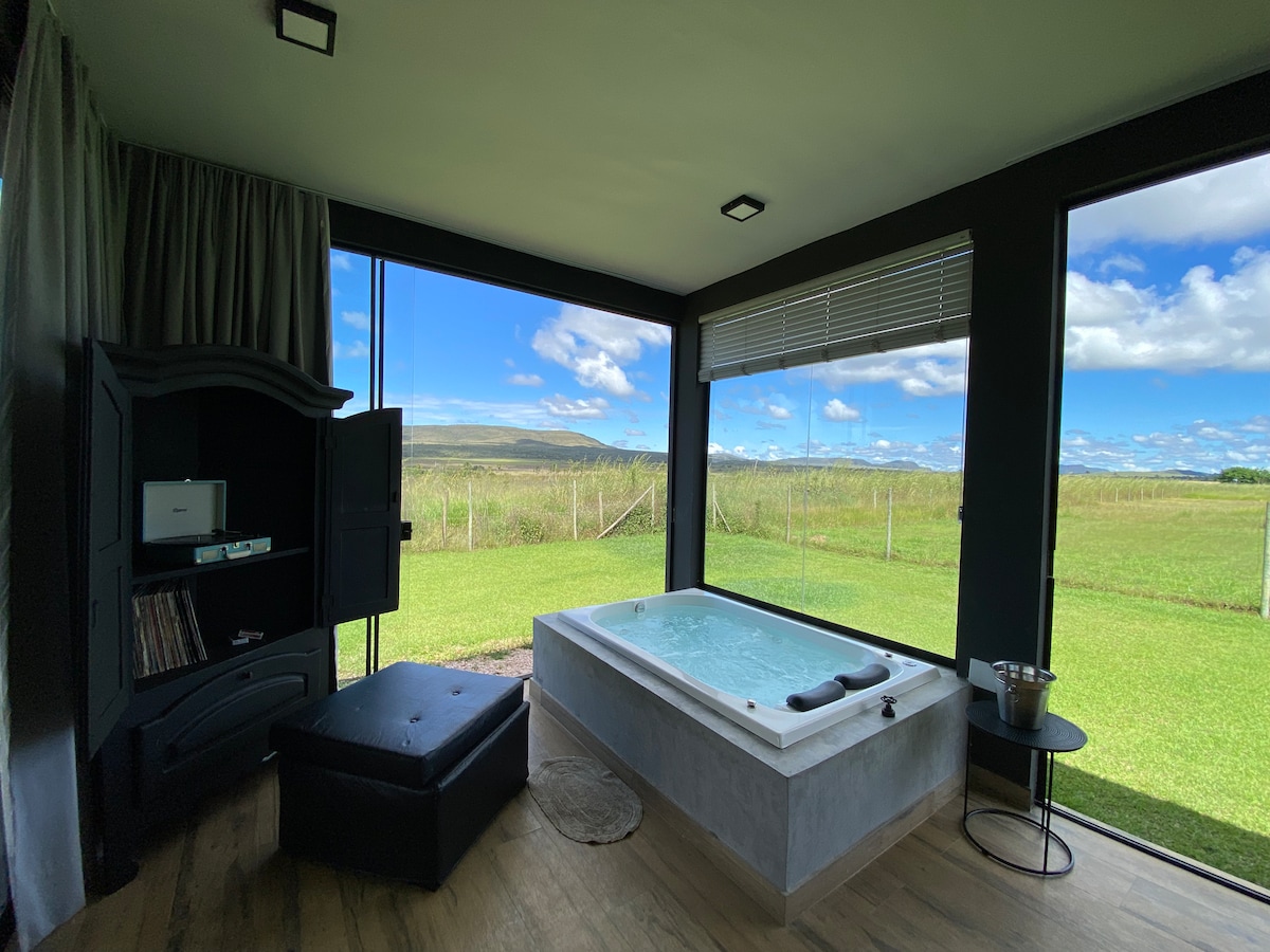 A spacious area is highlighted by a covered and heated jacuzzi, positioned next to large glass windows that frame expansive views of the surrounding greenery and mountains. A black ottoman and small table complete the relaxing ambiance, while soft curtains draw the eye.