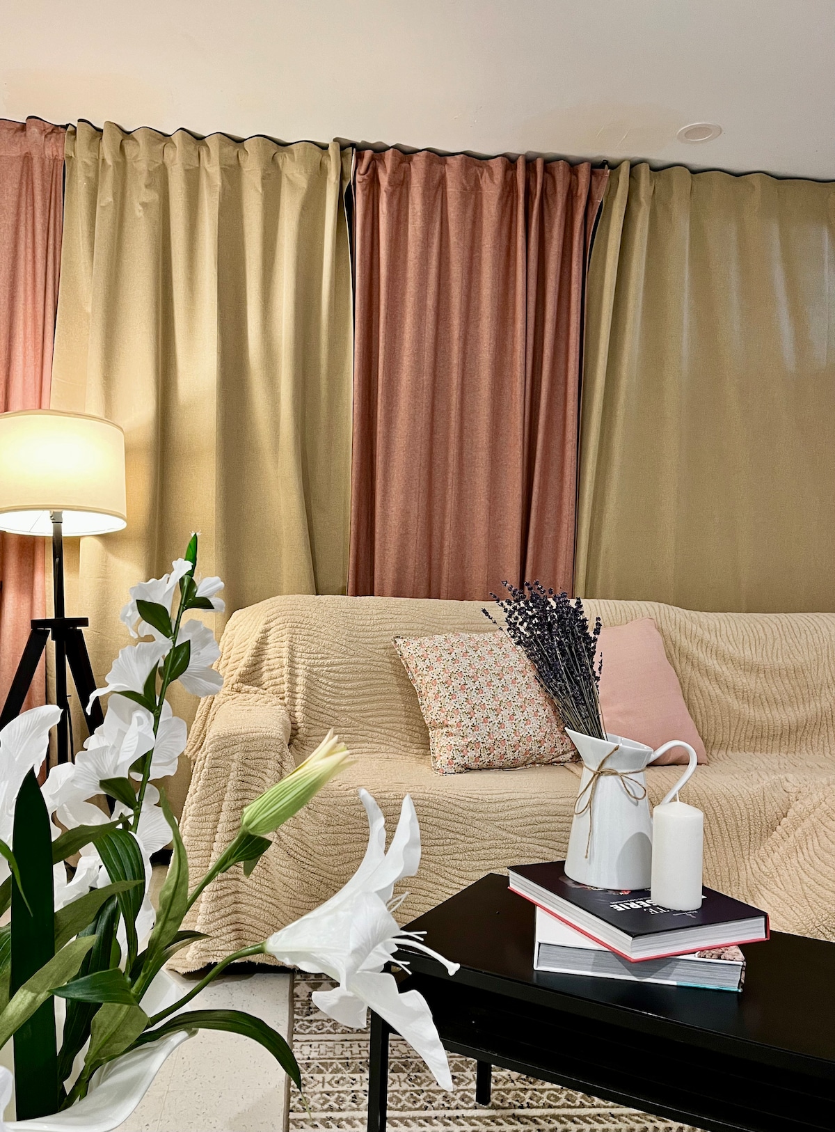 A living area features a beige sofa adorned with decorative cushions. Light-filtering curtains in soft tones add warmth to the space. A modern lamp stands beside the sofa, while a small black table displays a vase with flowers and a stack of books.