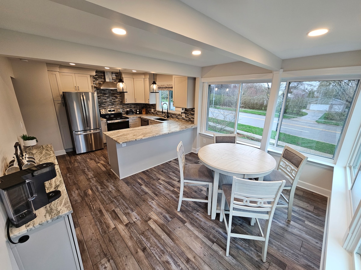 A modern kitchen and dining area feature light wood cabinetry and a spacious island with granite countertops. A round dining table is surrounded by four white chairs, while large windows offer natural light and views of the greenery outside.
