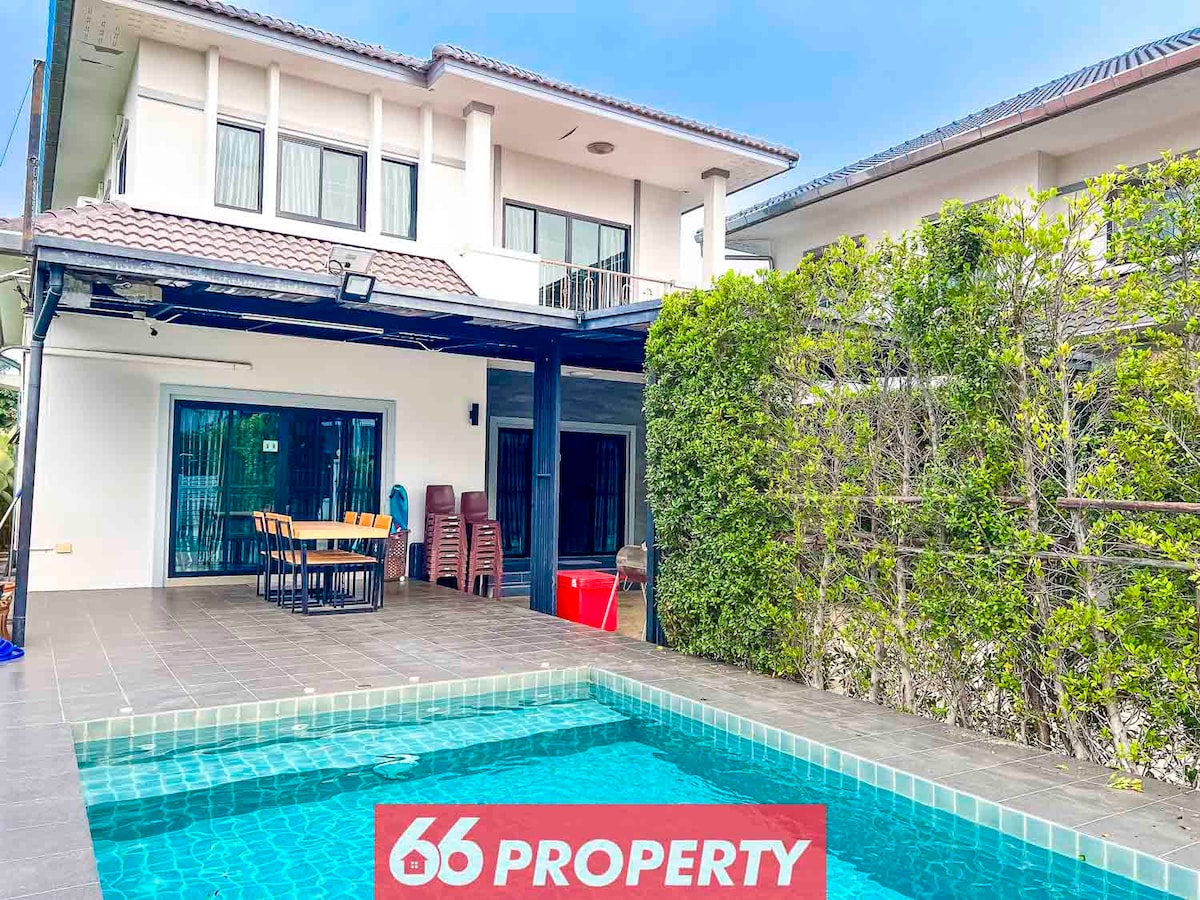 A private pool is set against a backdrop of a modern two-story house, featuring a spacious patio area with a dining table and chairs. Lush greenery frames the pool, providing a sense of privacy and tranquility. Large glass doors lead into the living space, enhancing the welcoming ambiance.
