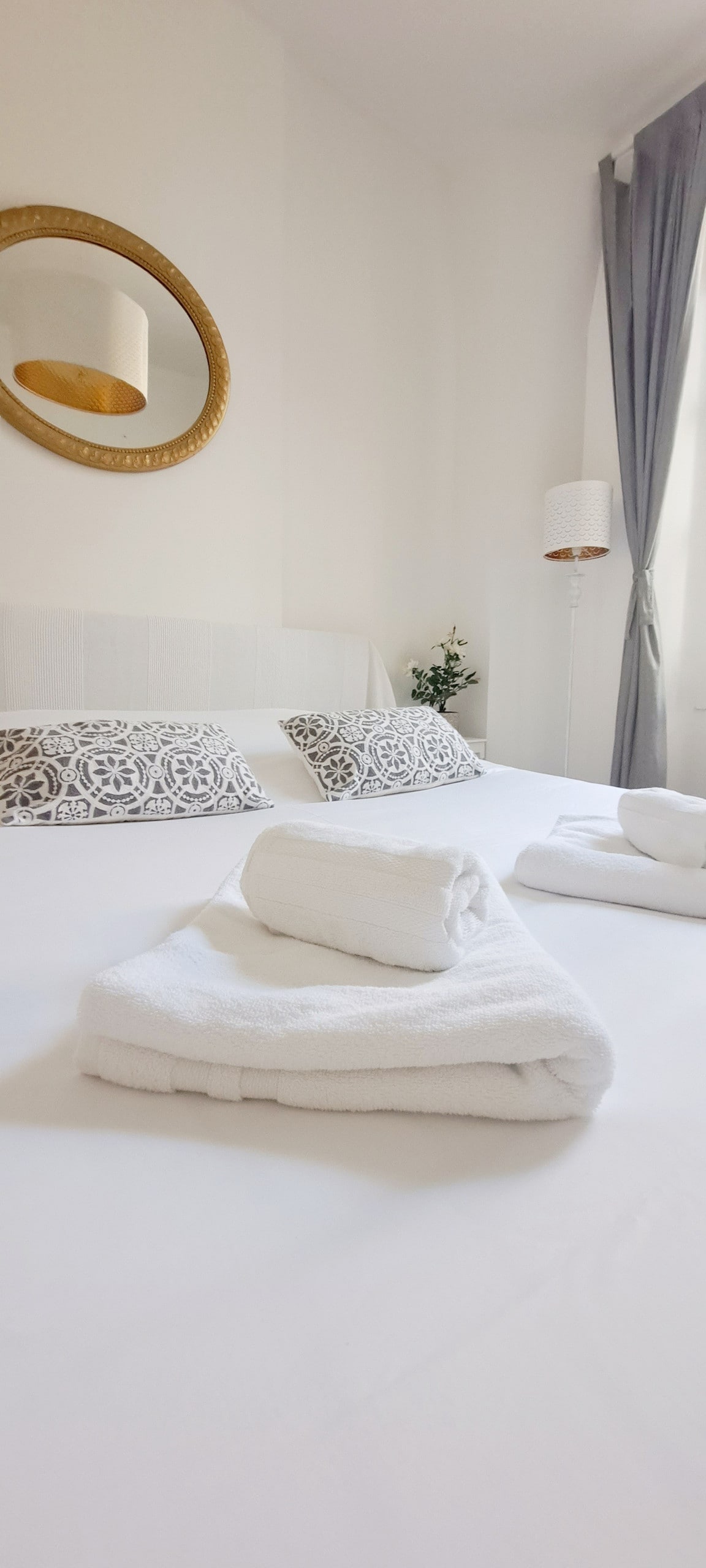 A bright bedroom is presented, featuring a king-size bed adorned with neutral bedding. Freshly folded towels are neatly arranged on the bed, complemented by decorative pillows. Natural light filters in through sheer curtains, highlighting the soft, calming tones of the space.