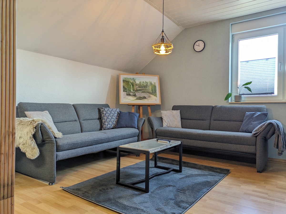 A cozy living area is presented, featuring two gray sofas adorned with decorative pillows. A coffee table rests on a soft rug, while a wall clock and a window provide functional elements. A framed painting of a landscape adds a decorative touch.