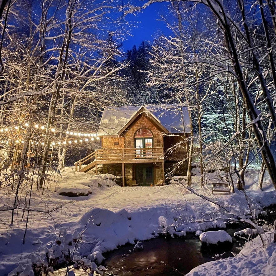 A cozy stone cottage is illuminated by soft string lights, surrounded by a serene winter landscape. Fresh snow blankets the ground, while the babbling brook is partially visible in the foreground, creating a tranquil and enchanting atmosphere.