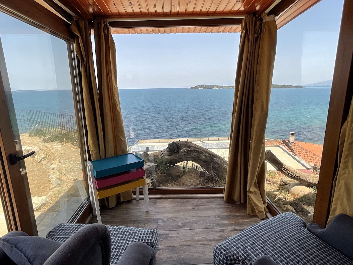 A inviting room features large glass windows providing panoramic views of the sea. Comfortable seating is positioned near the window, while a colorful small table adds a playful touch to the space. Natural light illuminates the wooden floor and warm curtains.