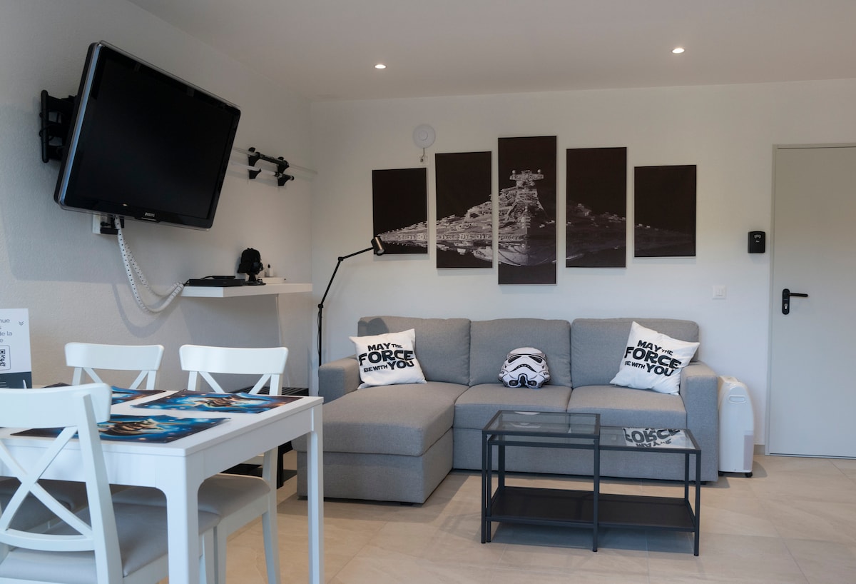 A modern living area features a gray sectional sofa positioned near a sleek coffee table. A wall-mounted television is visible, alongside a minimalist shelf. An art piece with black and white imagery enhances the decor. A dining table with white chairs completes the inviting space.