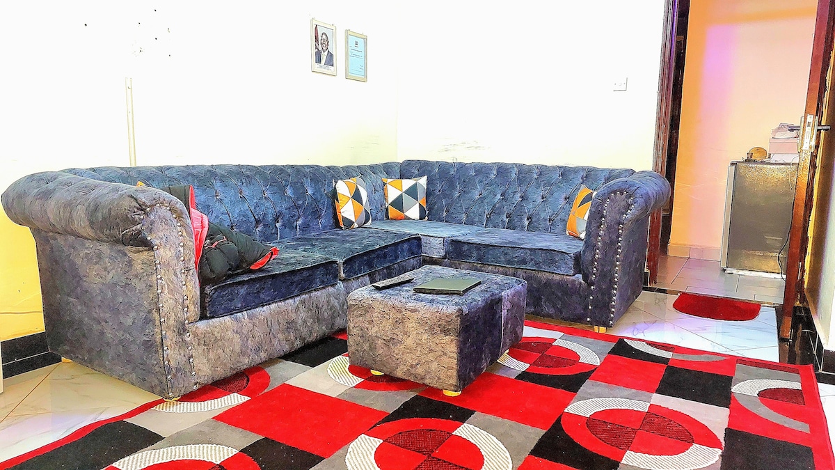A spacious living area features a large sectional couch in a deep blue fabric, accented by decorative pillows. A small ottoman sits in front of the couch on a vibrant area rug with geometric patterns. Soft lighting highlights the cozy setting.