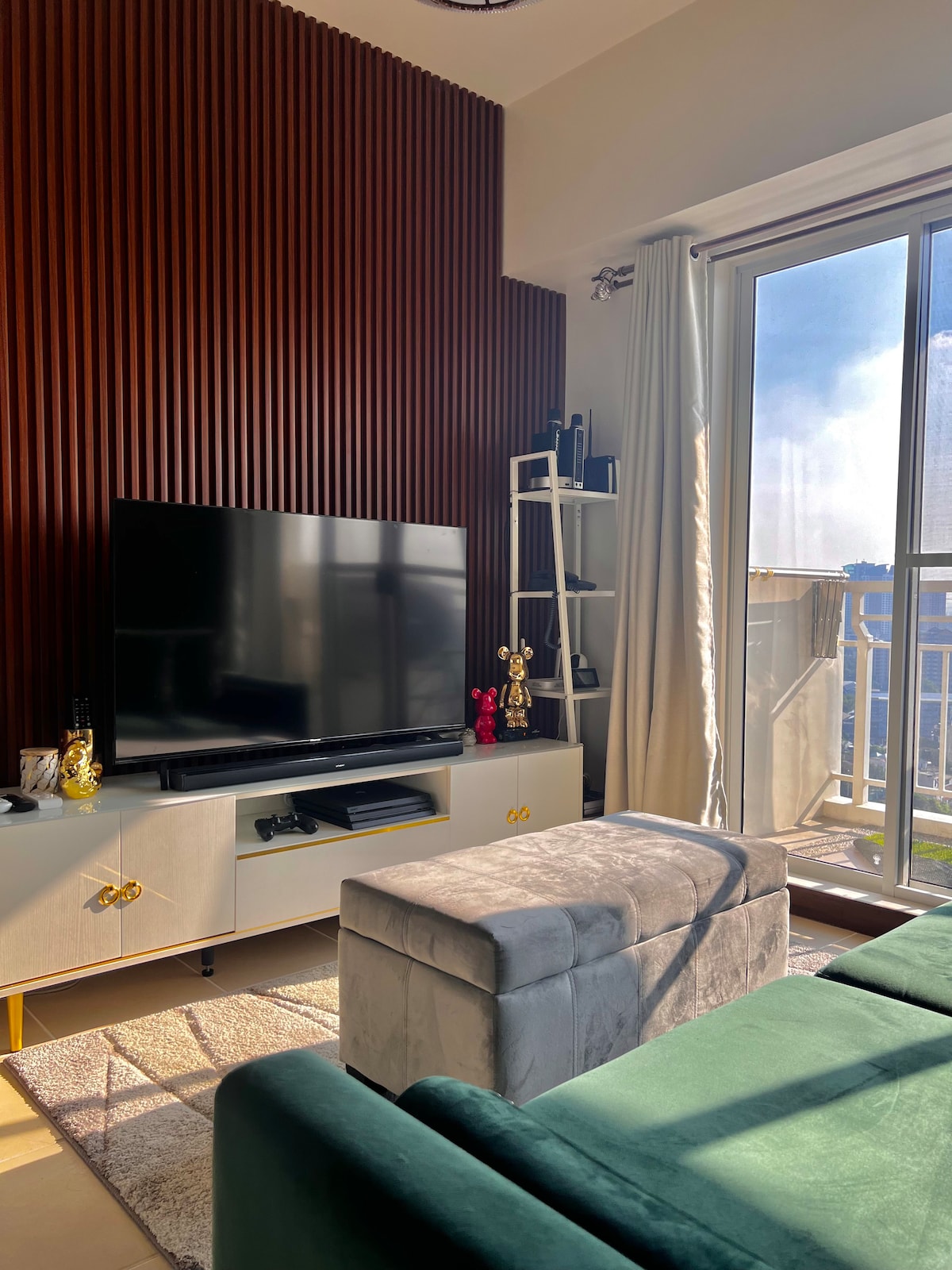 A modern living area is showcased, featuring a large flat-screen television positioned above a sleek console. Large windows provide ample natural light, while a compact ottoman offers seating. The warm, textured wall contrasts with the light furnishings, creating a balanced, inviting space.