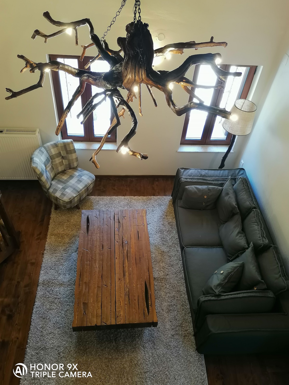 A spacious living area is illuminated by a unique chandelier made from natural materials. A large wooden coffee table is centered, surrounded by a comfortable sofa and a patterned armchair. Bright light filters through multiple windows lining the walls.
